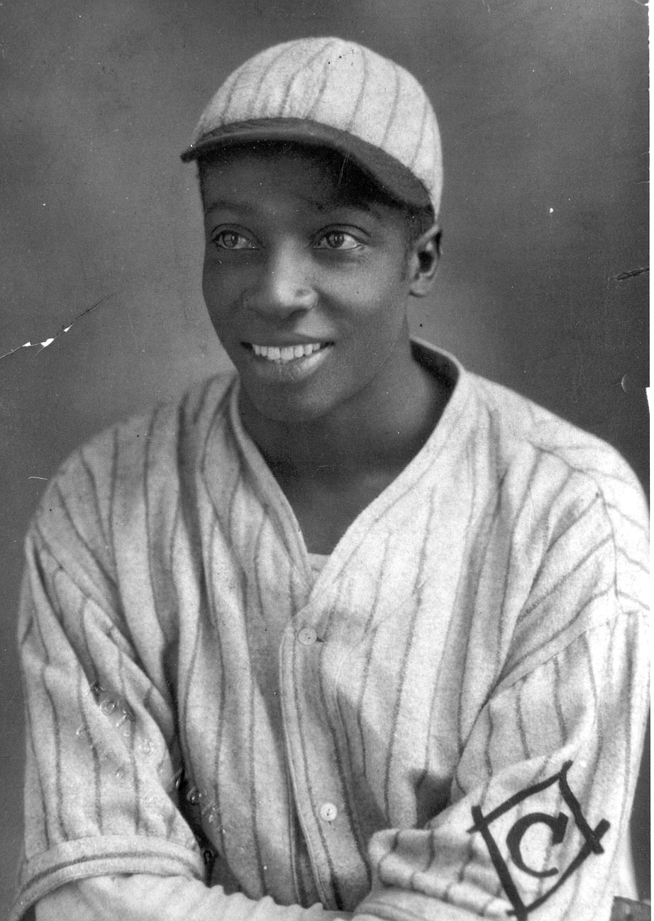 Smiling baseball player from the early 20th century, wearing a pinstripe uniform and cap. The jersey has an emblem on the left sleeve. The image is in black and white, showing a classic athletic pose and attire from that era.