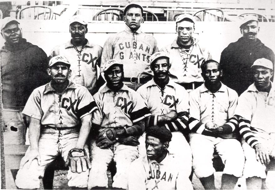 Black and white historical photo of a baseball team. Players are posing in two rows, wearing uniforms labeled Cuban Giants. The front row holds gloves. The background features empty wooden bleachers.