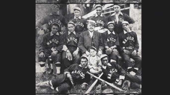 Historic black and white photo of a baseball team wearing uniforms labeled "Marico." The team poses with a man in a suit and cap and a young boy sitting in front. Bats are placed on the ground.