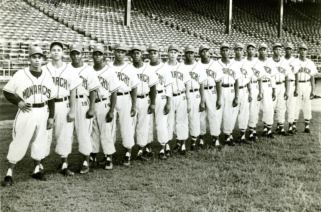 A vintage black and white photo of a baseball team lined up on a grass field. They wear uniforms with Monarchs written on the front, and baseball caps. The stadiums empty bleachers are visible in the background.