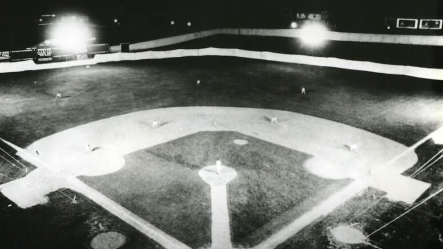 Black and white vintage photo, showing a bird's eye view of a baseball field at night, with lights in the background