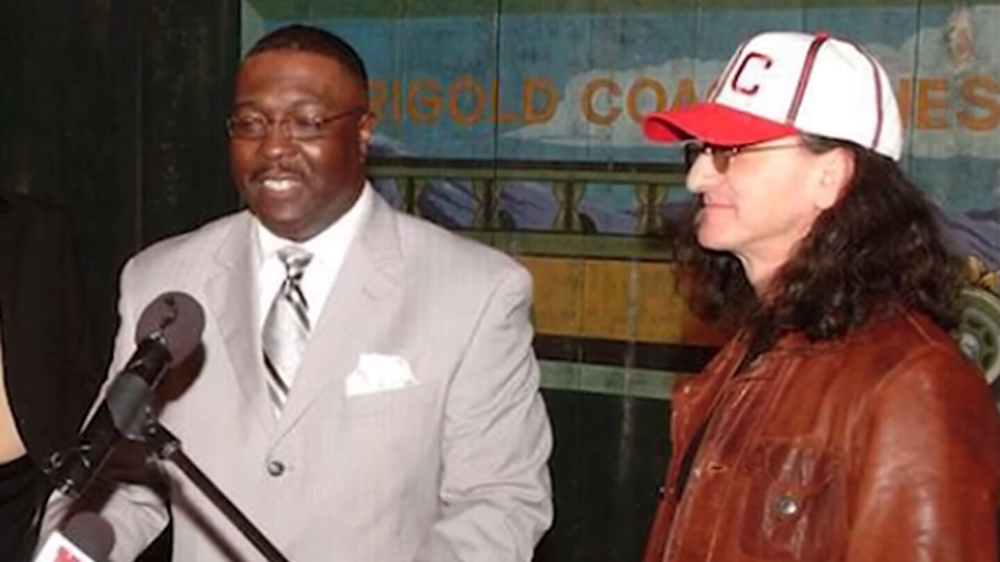 Two people standing together. The person on the left wears a gray suit and glasses, speaking into a microphone. The person on the right wears a baseball cap, brown jacket, and glasses. They are indoors with a colorful wall in the background.