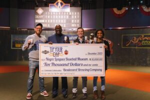 Four people stand smiling, holding bottles and presenting a large ceremonial check for $10,000 made out to the Negro Leagues Baseball Museum. They are indoors on a stage with a colorful backdrop and baseball-themed decor.