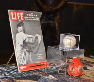 A vintage LIFE magazine with a baseball player on the cover is displayed next to a baseball in a clear case. A red strap or band is placed beneath the display on a patterned tablecloth.