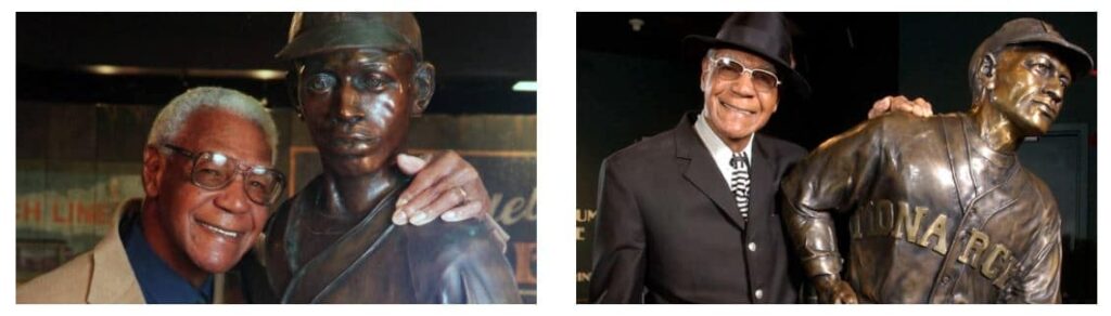 An elderly man smiles while standing next to two bronze statues. In the first image, the statue is of a baseball player wearing a cap. In the second, the statue is dressed in a baseball uniform with Monarchs written on it. The man wears a suit and hat.