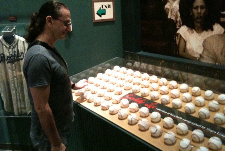 A person observes a display case of baseballs, each featuring signatures, in a museum setting. A vintage baseball jersey and an "Exit" sign are visible in the background.