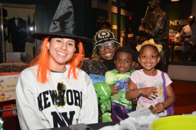 Four people in costumes, including a witch hat, glasses, Hulk, and a princess dress, smile at a table in a museum setting with "Negro Baseball" signs in the background.