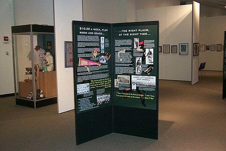 Museum exhibit with informational panels on baseball history, featuring text and historical images. Display case in the background with a baseball jersey and cap.