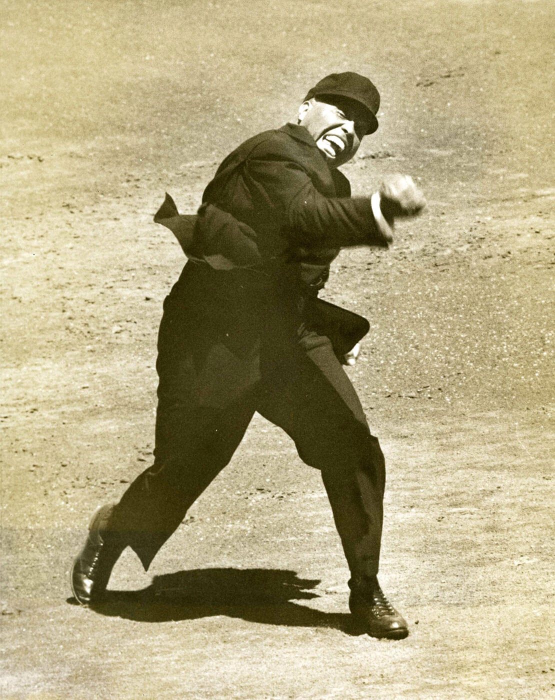 An umpire in a dark suit energetically gestures as he makes a call on a baseball field. He appears focused, with one arm extended and body slightly turned, capturing a moment of dynamic action. The ground is an earthy, textured surface.