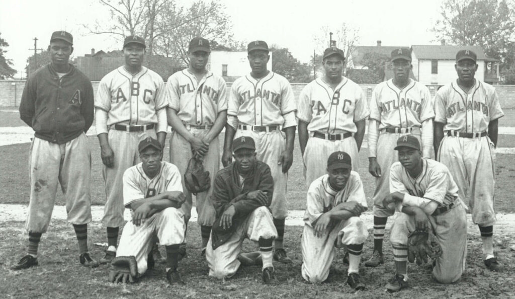 A vintage black and white photo of a baseball team with eleven players. Six players are standing in the back row wearing uniforms with ABC and Atlanta visible. Five players are kneeling in the front row. Trees and buildings are in the background.