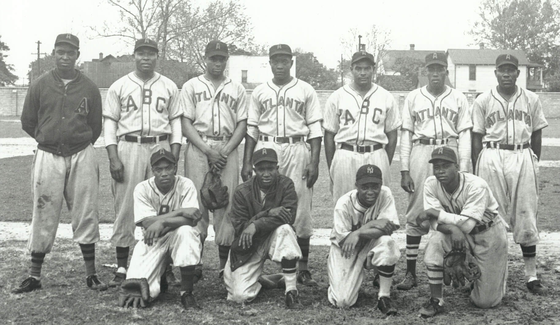 A vintage black and white photo of a baseball team with eleven players. Six players are standing in the back row wearing uniforms with ABC and Atlanta visible. Five players are kneeling in the front row. Trees and buildings are in the background.
