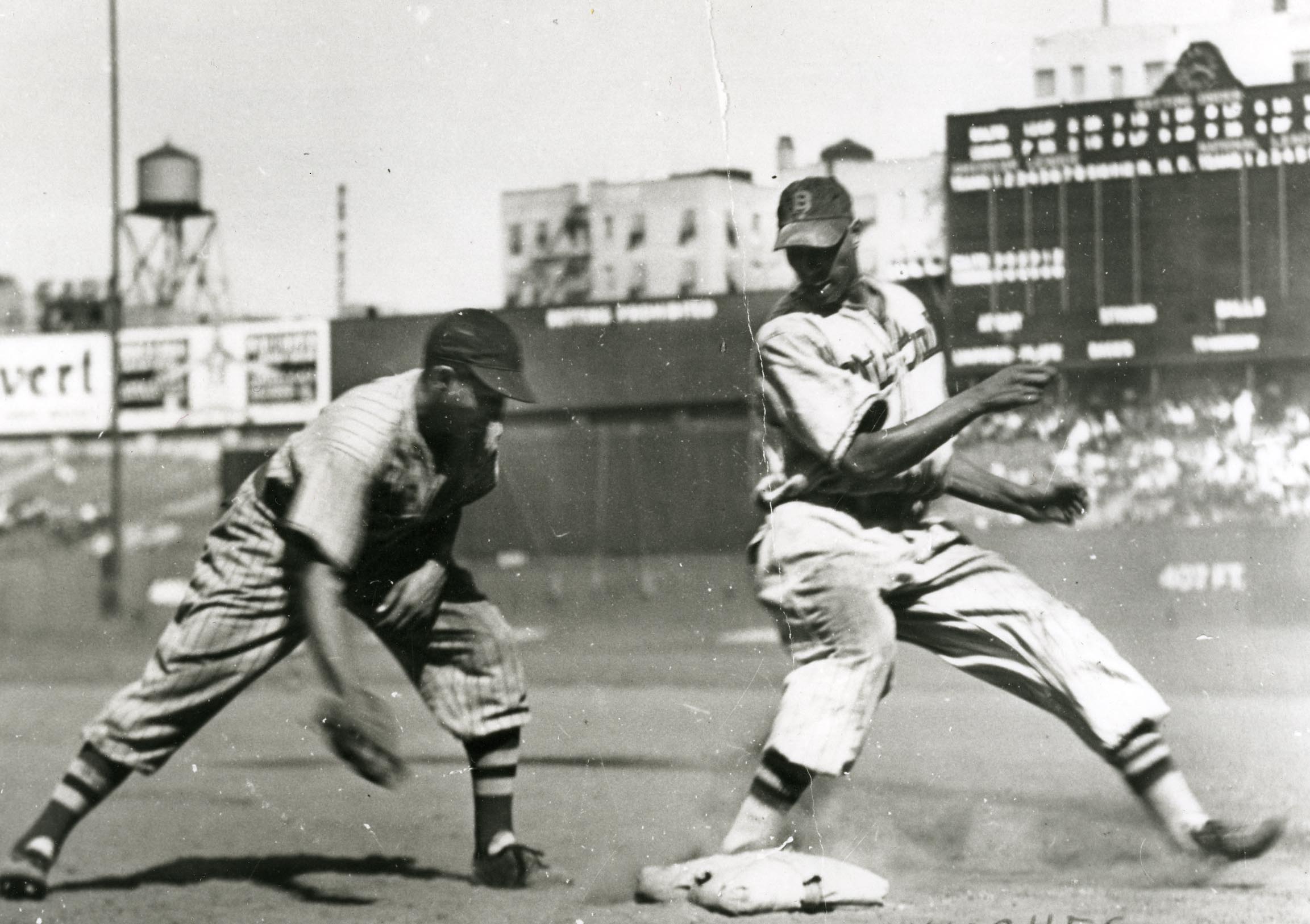 Two baseball players in vintage uniforms during a game. The runner is sliding into a base, while the other player, in a striped uniform, reaches to tag. A scoreboard and buildings are visible in the background.