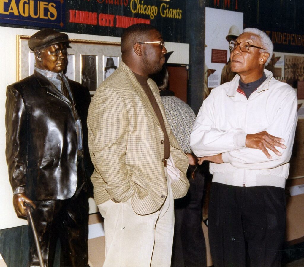 Two men converse in a museum, with one wearing a beige jacket and glasses and the other in a white jacket with arms crossed. They stand next to a bronze statue of a man in a suit and cap. Baseball memorabilia is visible in the background.