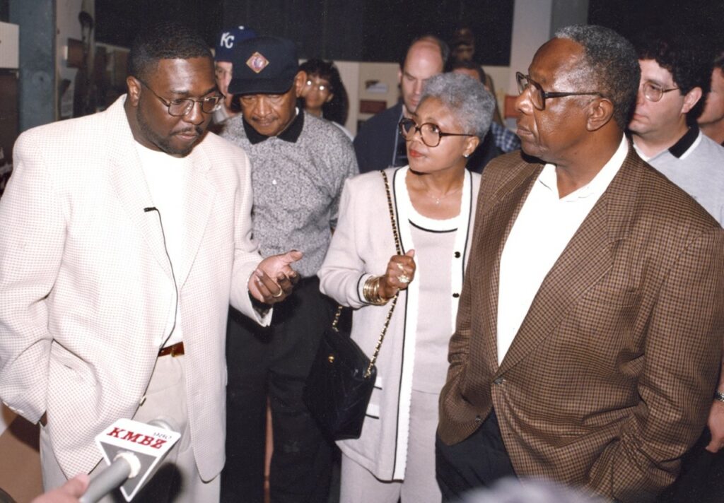 A group of people are engaged in conversation indoors. A man on the left gestures as he speaks, while others listen. A person in the background wears a cap with a logo. A microphone with a KMBC label is seen in the foreground.