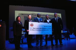 Five people stand on a dark stage holding a large check for the Negro Leagues Baseball Museum, dated November 11, 2022. They are smiling, and the background features a projected screen with partially visible text.
