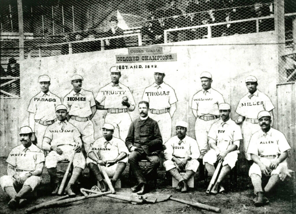 A vintage black and white photograph of a baseball team with twelve players in uniforms and a manager seated in the center. The sign above reads Colored Champions. 1887 and 1888. Names are visible on the players shirts.