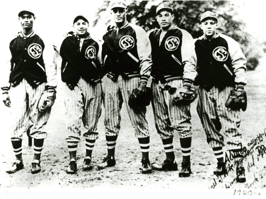 Black and white photo of five young baseball players standing side by side. They are wearing striped uniforms and jackets with a CS logo. Each holds a baseball glove. Trees are visible in the background.