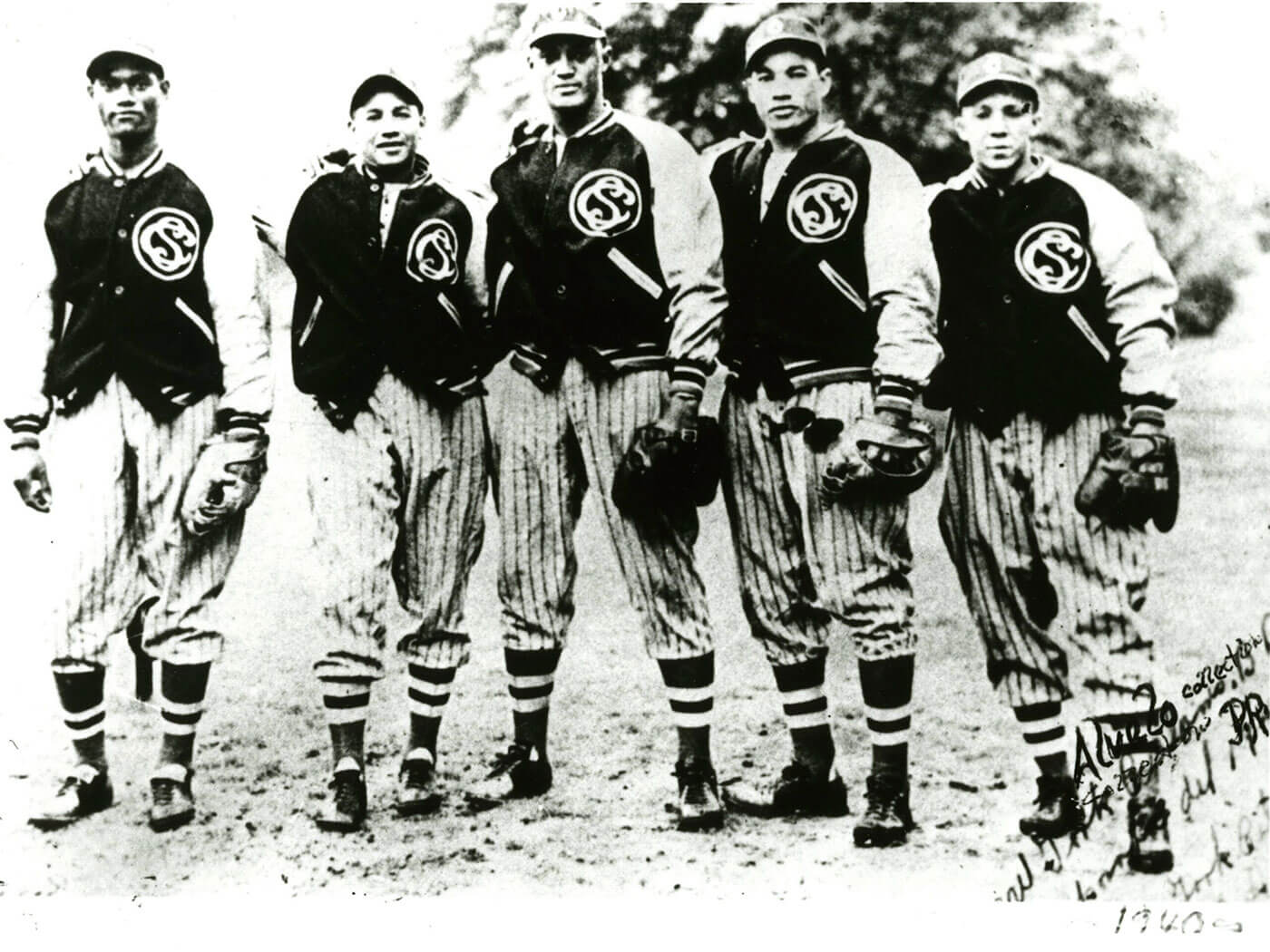 Black and white photo of five young baseball players standing side by side. They are wearing striped uniforms and jackets with a CS logo. Each holds a baseball glove. Trees are visible in the background.