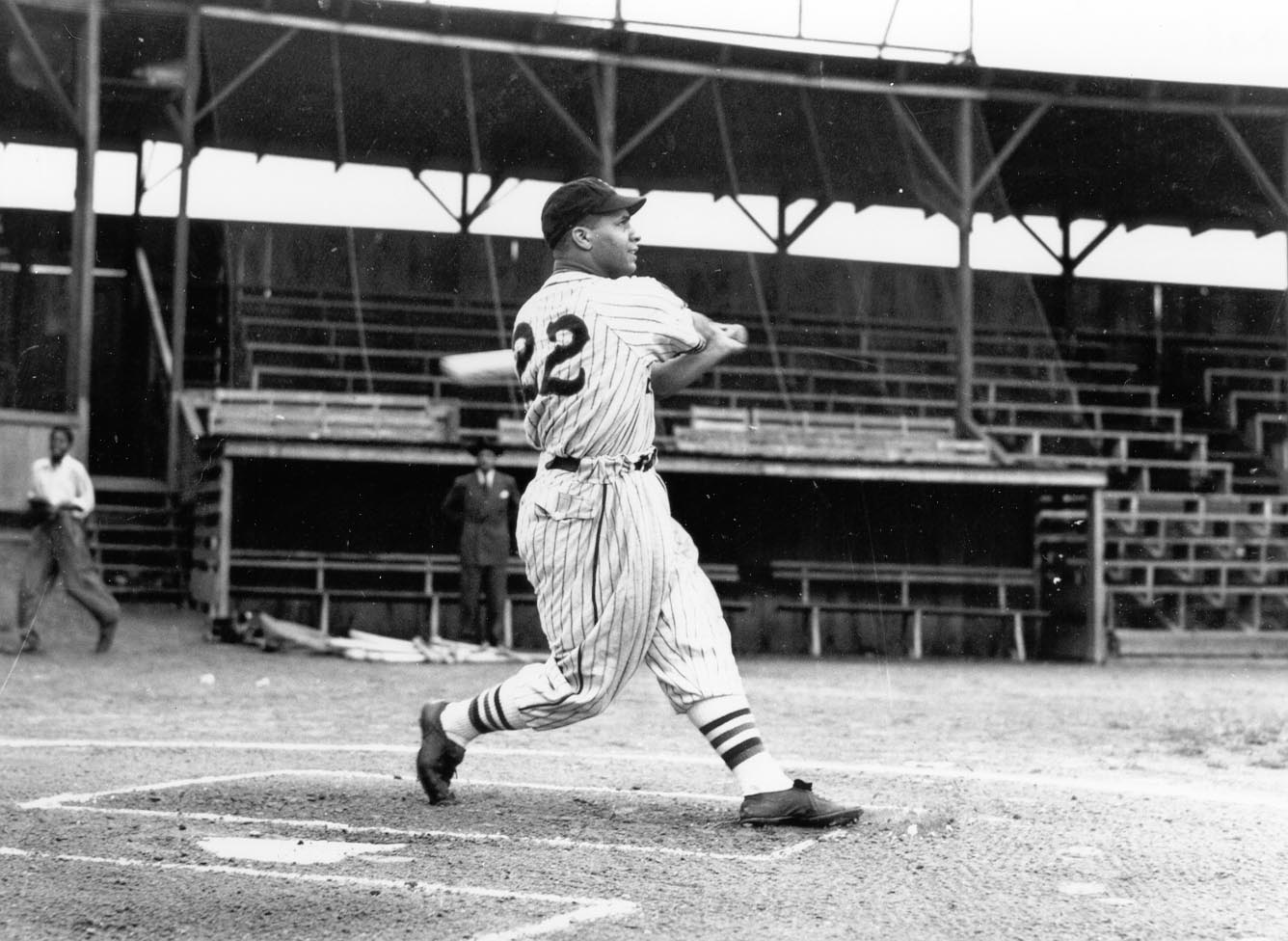A baseball player wearing a striped uniform with the number 23 swings a bat on a baseball field. He appears mid-swing, with empty stadium seating and a few onlookers in the background.