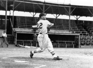 Black and white photo of a baseball player wearing a uniform with the number 22, swinging a bat. The scene is set in an empty stadium with bleachers in the background.