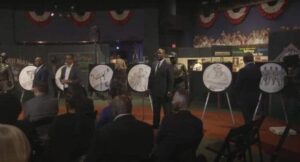 People in formal attire stand in a room with several round displays featuring art. The setting appears to be a museum or exhibit with informational displays and statues.