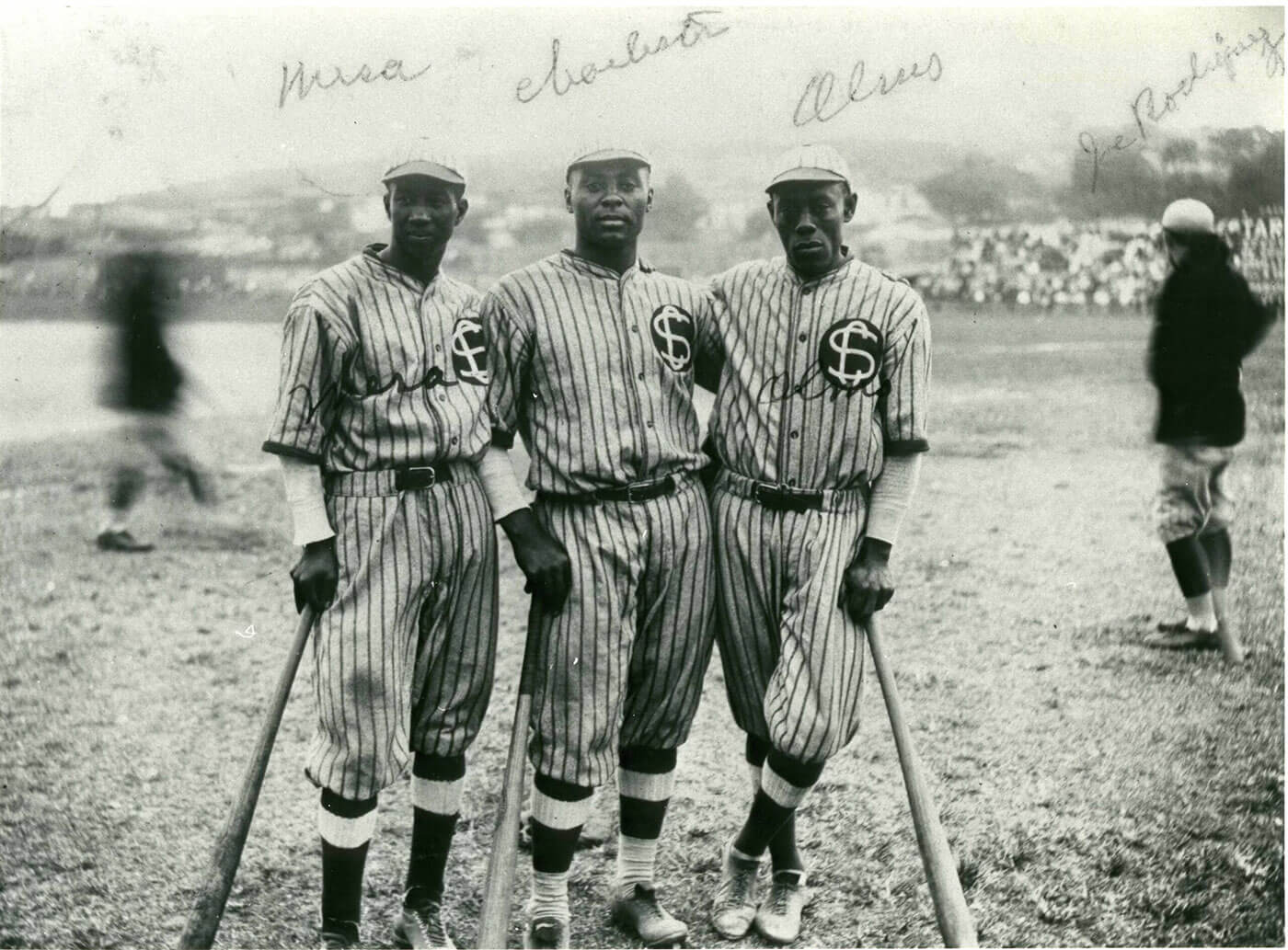 Three baseball players in striped uniforms stand on a field holding bats. They are part of a historic team, with a blurred crowd in the background. The image is black and white, and names are handwritten above them.