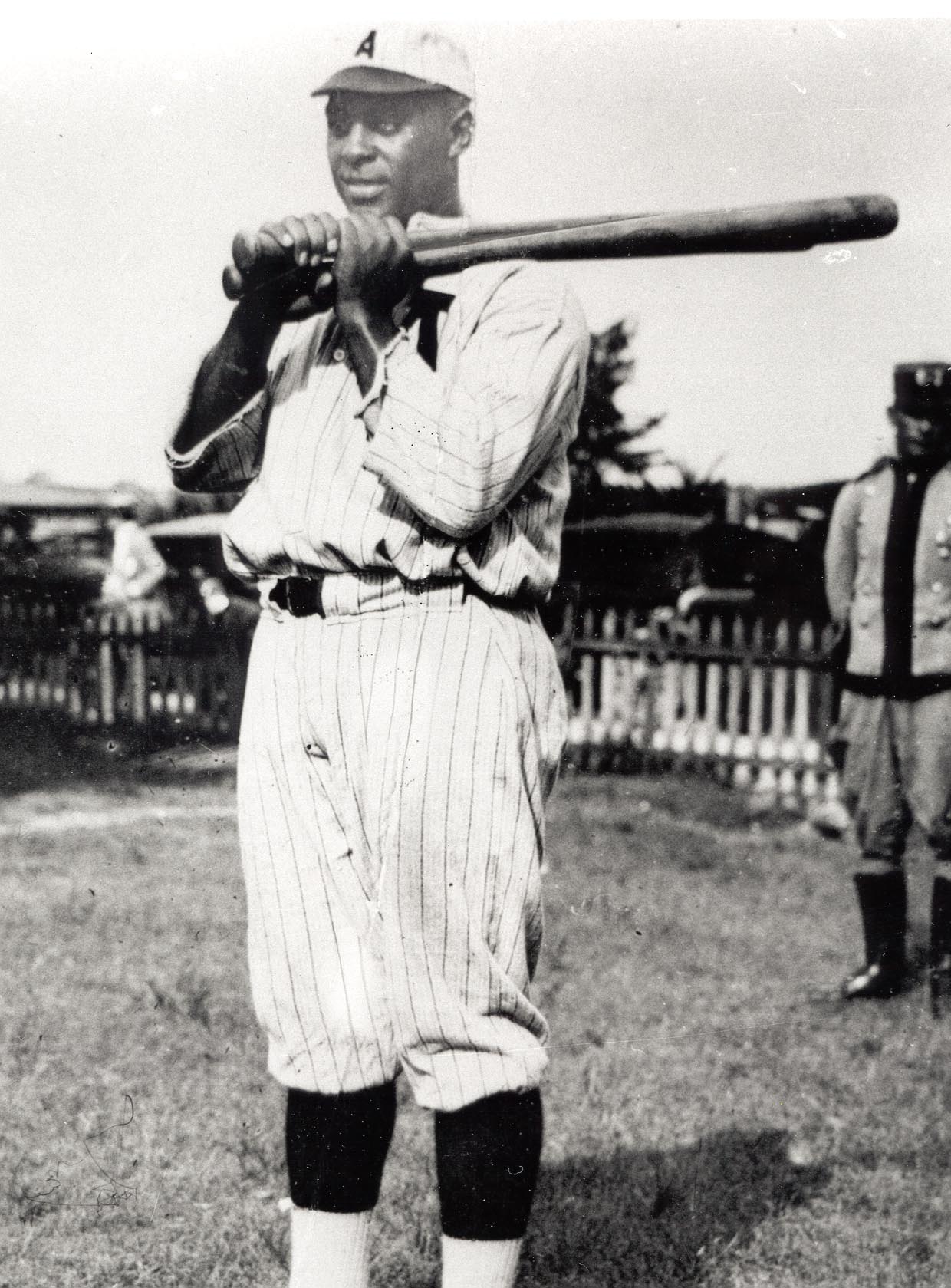 Black and white photo of a baseball player in a vintage uniform with vertical stripes, holding a bat over his shoulder. He stands on a grassy field with a fence and people in the background.