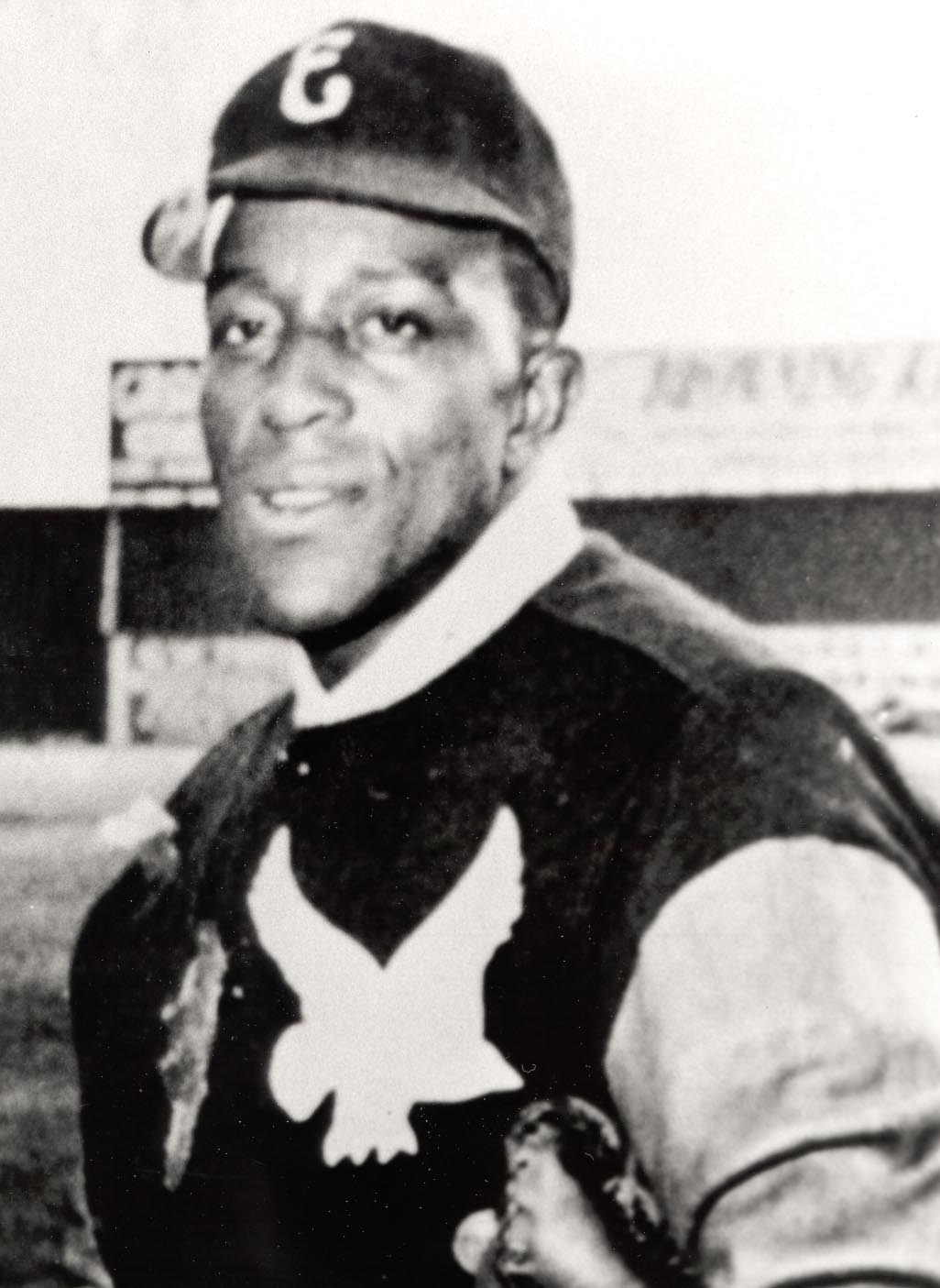 A baseball player wearing a cap and jersey with an eagle emblem is holding a glove. The background features a blurred field and billboard. The image is black and white.