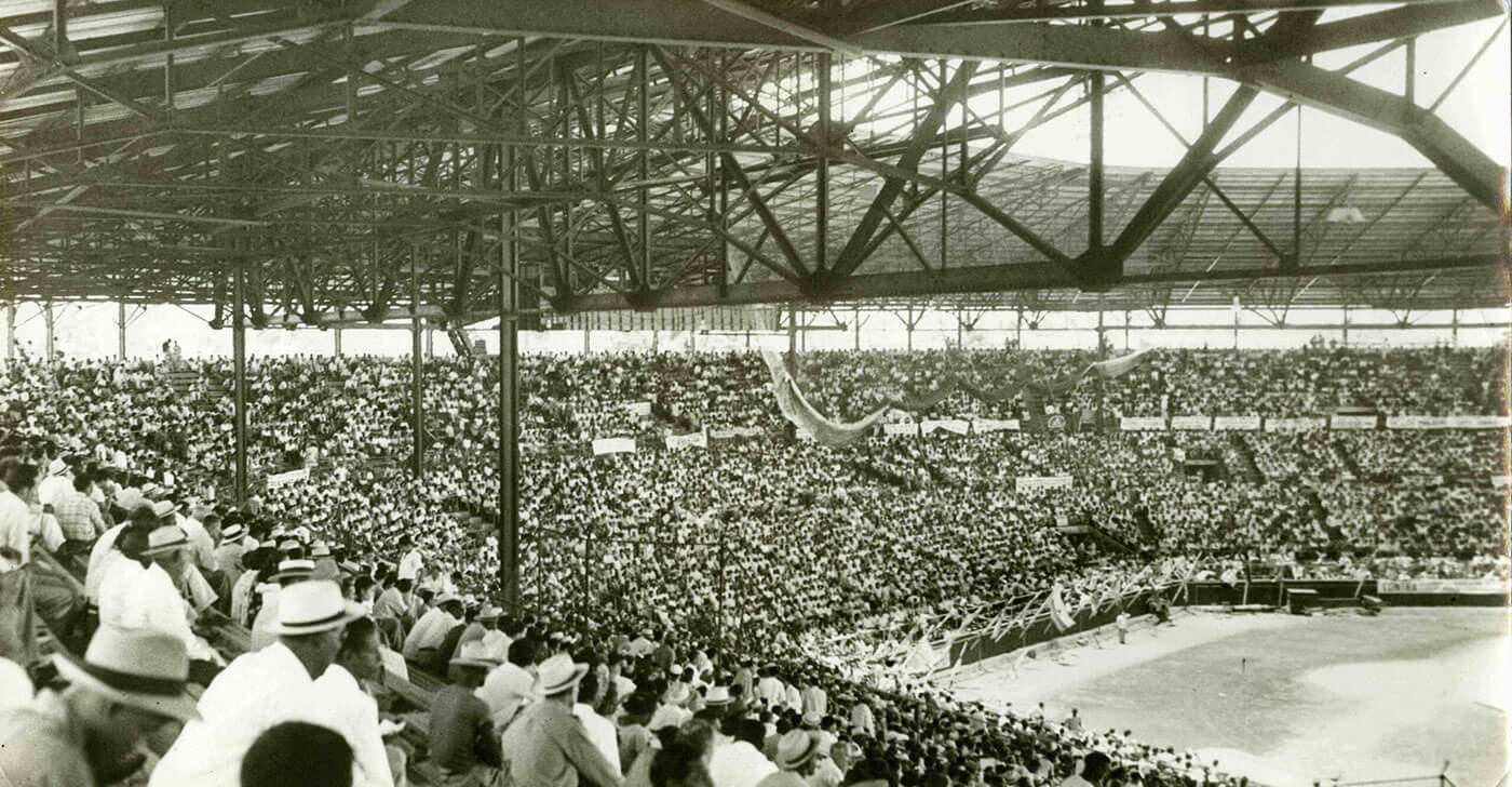 A black and white photo of a packed baseball stadium. The crowd fills the stands, many wearing hats. The field is visible with banners hanging around. The atmosphere seems lively and engaged in the event.