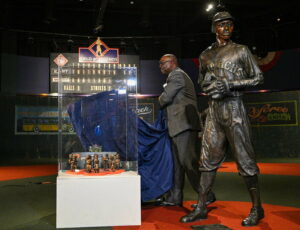 A man unveils a glass display case containing miniature baseball statues. Next to him stands a life-sized statue of a baseball player in a uniform labeled Monarch. The background features vintage baseball-themed decor.