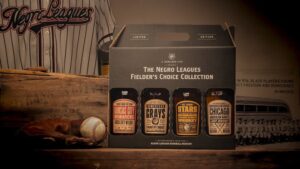 A limited edition box set of five whiskey bottles, each labeled with names of Negro Leagues baseball teams. The background features a vintage baseball jersey, bat, glove, ball, and a historic photograph. Text mentions Negro Leagues Baseball Museum.