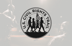 Logo of the U.S. Civil Rights Trail featuring silhouettes of three people walking, one holding an American flag. The background is a grayscale, blurred street scene.