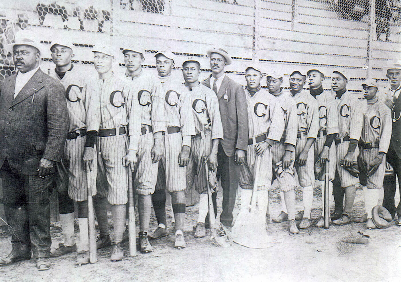Historic black and white photo of an early 20th-century baseball team, consisting of African American players in striped uniforms and caps, holding bats. They are lined up in front of a wooden fence, accompanied by two men in suits.