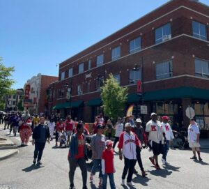 A group of people wearing red and white baseball jerseys and caps walk down a sunny street in front of a large brick building. Banners and a marquee sign are visible in the background.