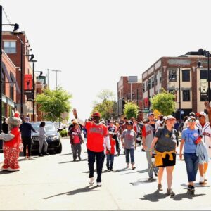 A crowd of people walks down a sunny street, some wearing red and white baseball jerseys. Buildings line the street, and trees provide shade. One person waves while others carry backpacks. The atmosphere is lively and festive.