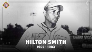 Black and white photo of a baseball player in a uniform with KC on it, wearing a cap. The text Hilton Smith 1907-1983 is at the bottom of the image. A vintage logo is in the top left corner.