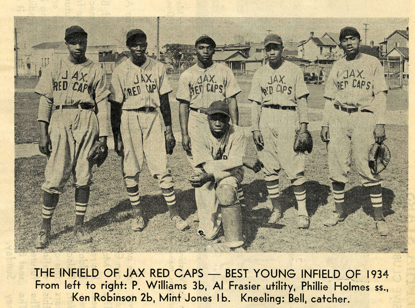 A vintage black-and-white photo of five baseball players from the Jax Red Caps, a team in 1934. Four players stand while one kneels, all wearing Jax Red Caps uniforms with caps. A residential neighborhood is visible in the background.