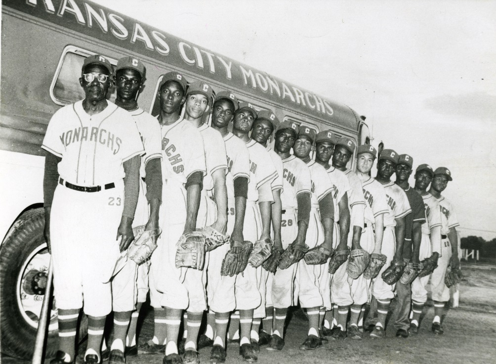 A black and white photo of the Kansas City Monarchs baseball team in uniform, standing in a line beside a bus. They are wearing hats and gloves, with Monarchs visible on their jerseys. The bus has the teams name on it.