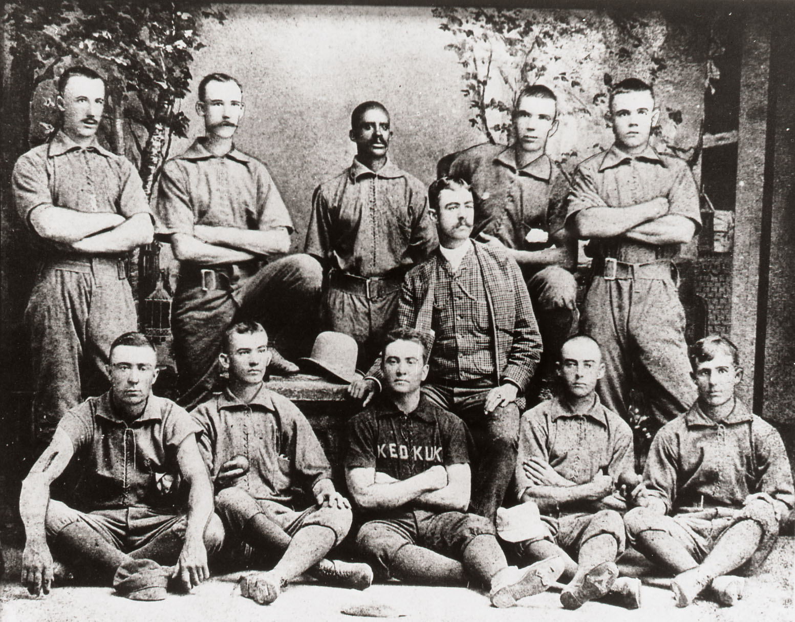 A vintage black and white photograph of a baseball team. Eleven men pose in uniforms, some standing and others sitting. Most have mustaches, and one wears a jersey with Keokuk on it. The background features a painted scenic backdrop.