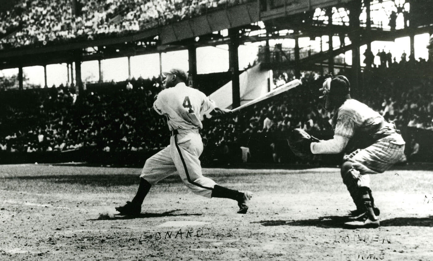 A baseball player wearing a uniform with the number 4 is swinging a bat on a field. A catcher is crouched behind him, wearing protective gear. The stadium is filled with spectators. The image is black and white, capturing a classic baseball scene.