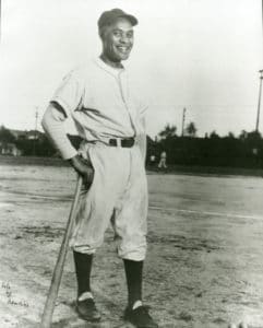 A vintage black and white photo of a baseball player standing on a field, wearing a classic uniform with a cap, holding a bat. The image captures the player smiling, with trees and some distant structures in the background.