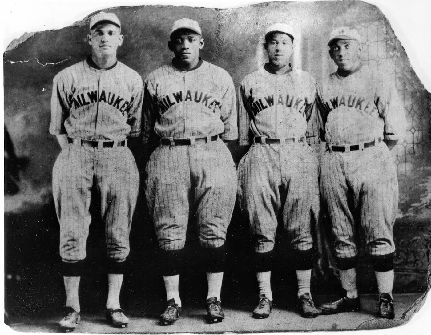 Four baseball players in vintage uniforms stand in a row. Their jerseys read Milwaukee. The photo is in black and white, with a rustic backdrop. They wear caps and high socks typical of early 20th-century baseball attire.