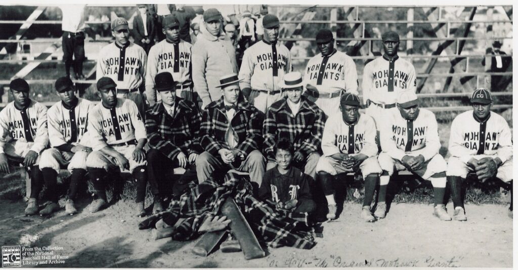 Black and white photo of the Mohawk Giants baseball team. Players in matching uniforms sit and stand in rows with a few men and a child in plaid jackets at the front. They pose outdoors near a wooden fence.