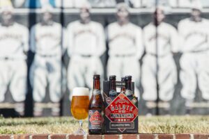 A six-pack of Boulevard Brewing Co.s Salute to the Monarchs beer is on display, with one bottle and a filled glass beside it. The background features a black and white mural of a historical baseball team.