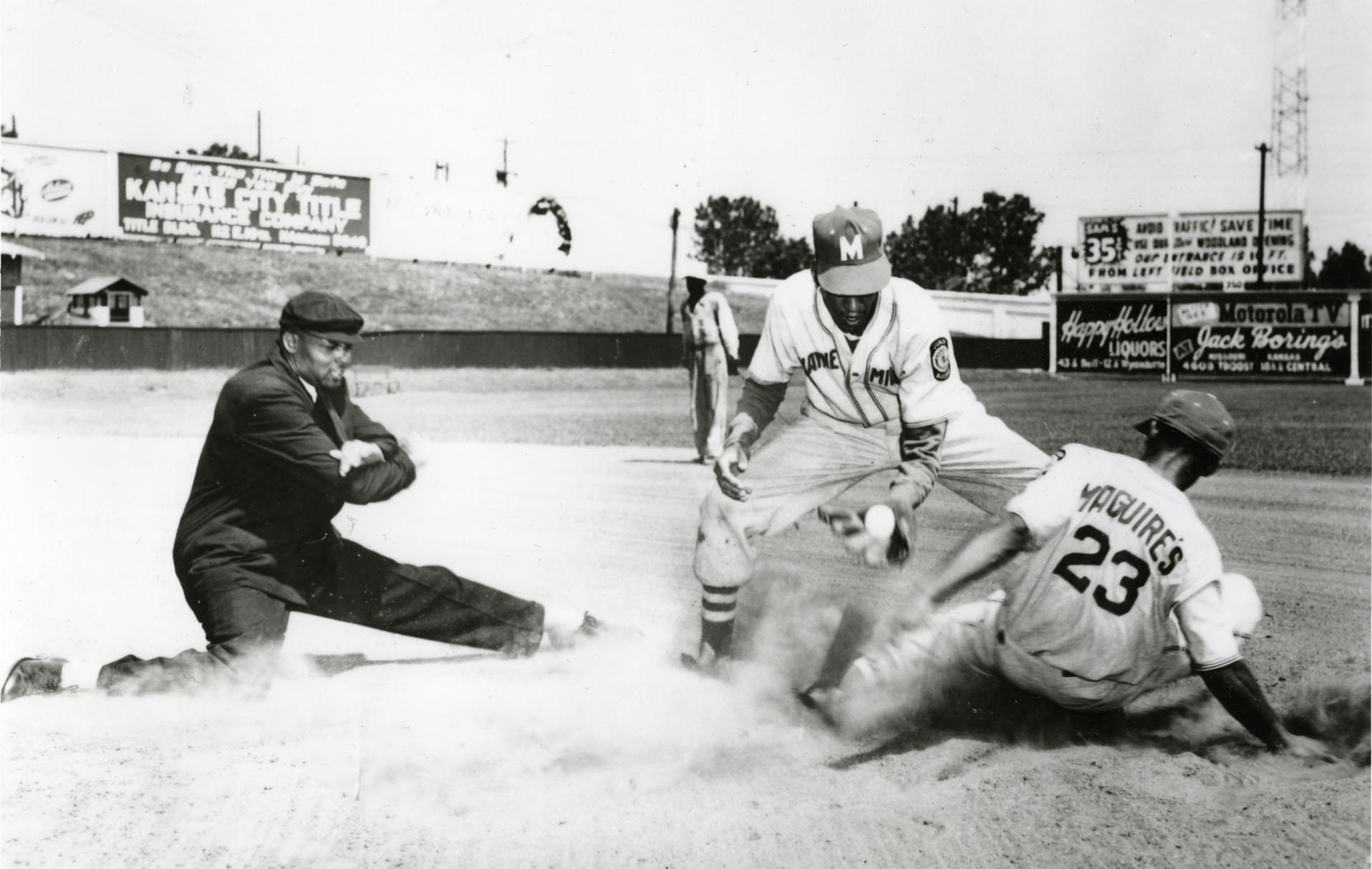 A baseball player sliding into a base, wearing a jersey with the number 23. An umpire is crouched, making a call, while another player stands nearby, wearing a cap with the letter M. Dust is rising from the slide. Billboards are visible in the background.