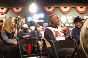 A group of people sit attentively in a room decorated with sports memorabilia and bunting. A man in the center wears a white and red sports jersey. Some attendees wear caps, and cameras are set up at the back. The atmosphere is focused and engaging.