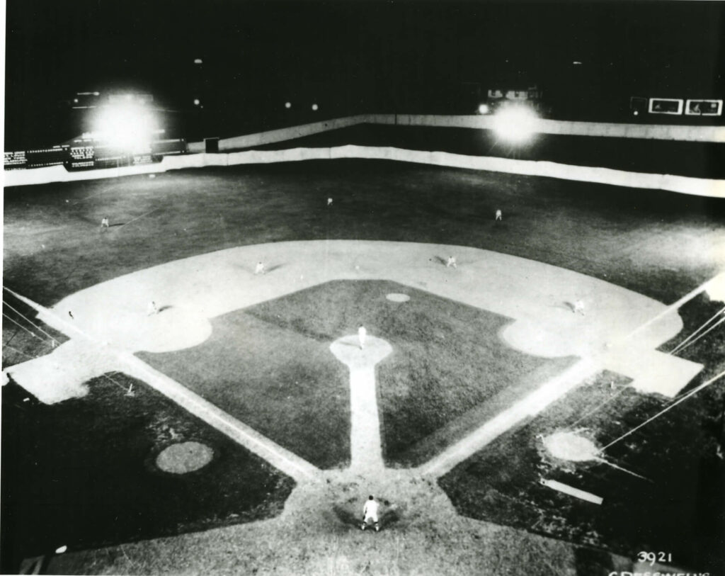 A black and white aerial photo of a baseball field under bright lights at night. Players are positioned on the diamond and outfield, with bases visible. The stands are empty, emphasizing the fields symmetry and lighting.