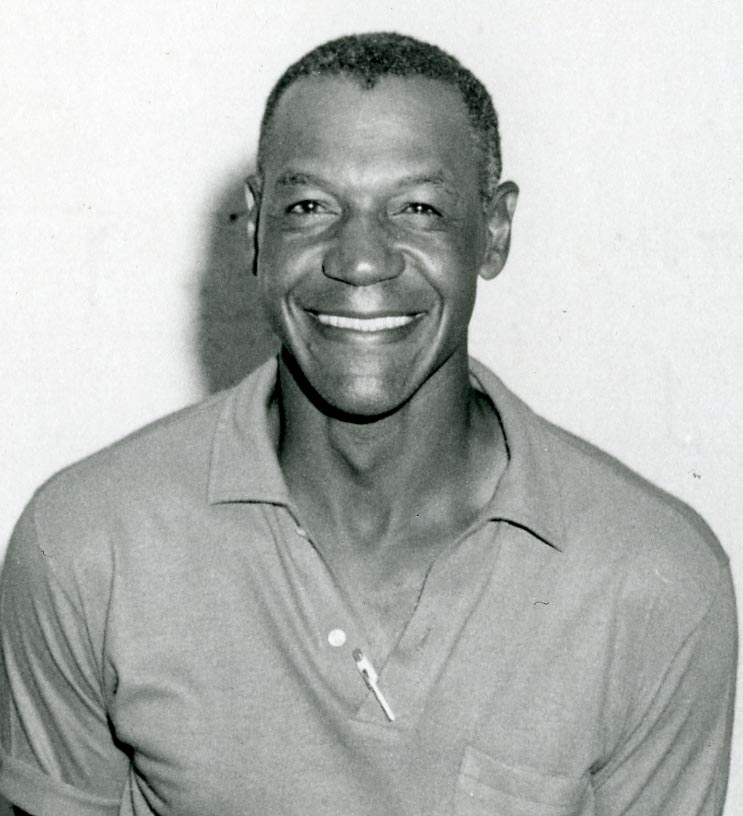 Smiling man wearing a short-sleeved, button-up shirt. The background is plain and light-colored.