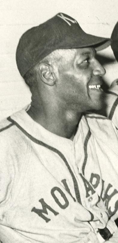 Smiling baseball player in a vintage uniform and cap, featuring Monarchs across the chest. The image is in black and white, capturing a candid side profile.
