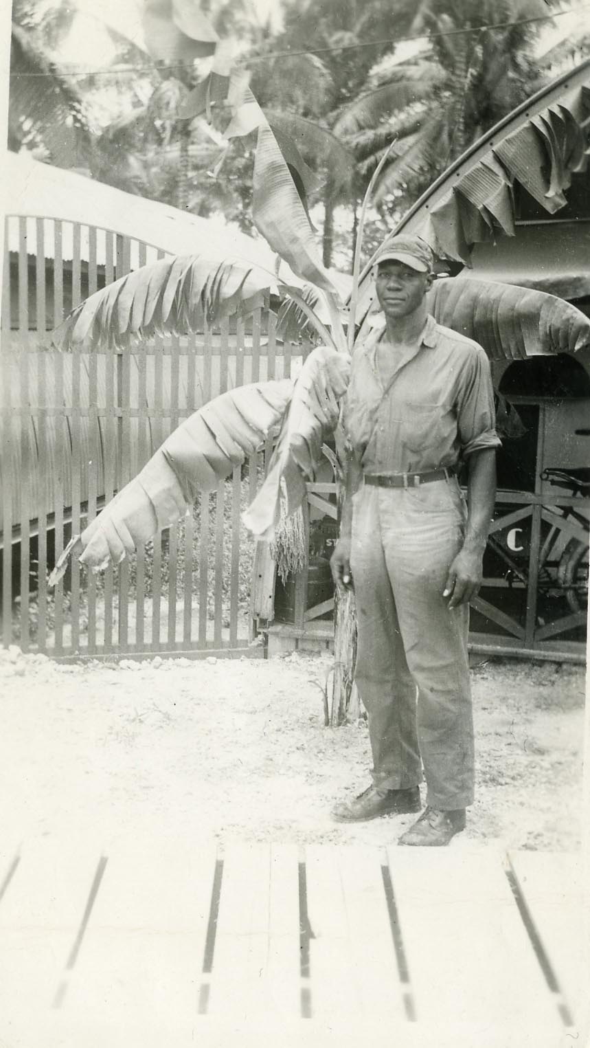 A man in a military uniform stands outdoors on a wooden walkway. Behind him are banana plants and a fence. The setting appears tropical.